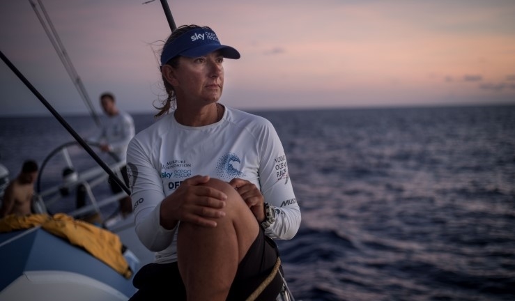 Dee Caffari in white t-shirt and blue cap against background of sea and sunset.