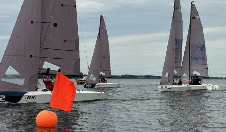 Four teams aboard RS21s racing at Rutland on a grey day in the British Keelboat League with an orange buoy and flag at the pin end and committee boat in the background. 