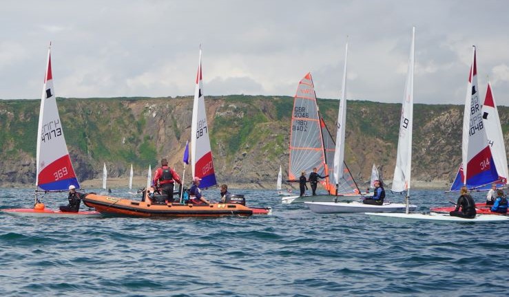 Diverse mix of youth and junior boats with colourful sails gathered around a RIB at the Regional Junior Championships for Wales 2023.
