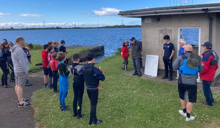 Youngsters gathered around for a briefing with race officers/coaches and white board outside next to the clubhouse at Tata Steel SC, ready for Dragon Series event 2024.