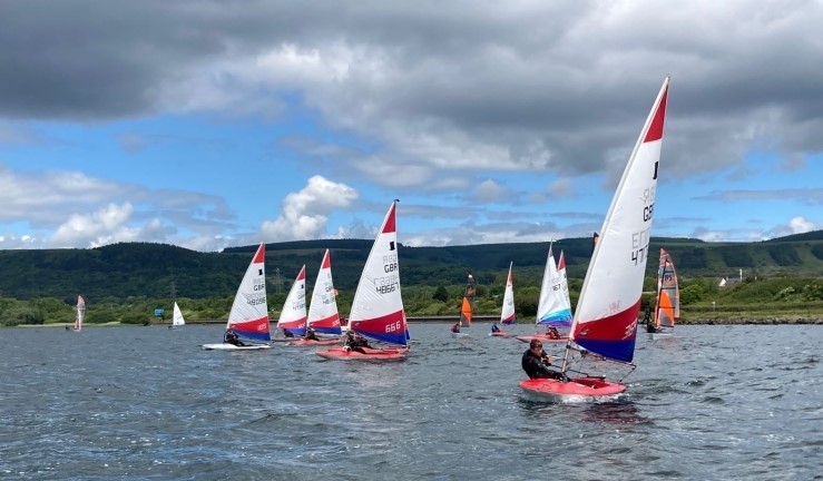 Topper dinghies racing upwind in the foreground with different junior classes behind against backdrop of hills and cloudy/blue sky.