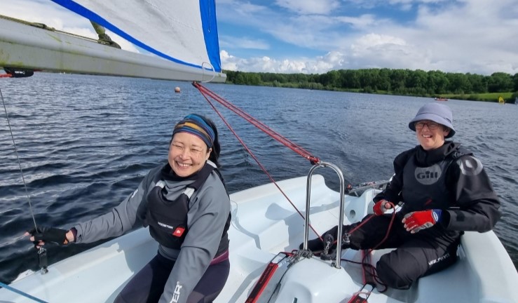 Two women smiling as they sail a dinghy on a sunny day at Burton SC as part of an RYA Midlands women's development day for aspiring female instructors.