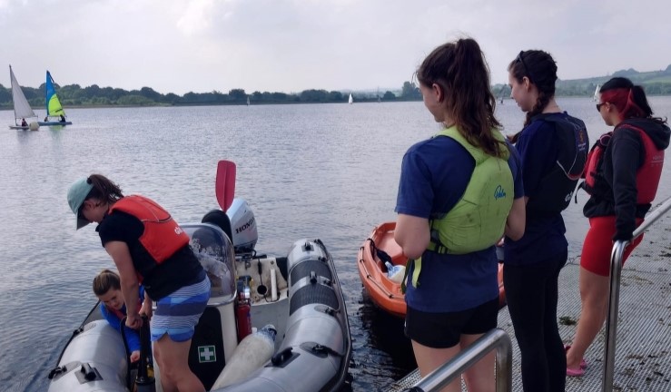 Three women on shore and waiting to get in a powerboat and two on the water in a grey RIB getting ready to go.