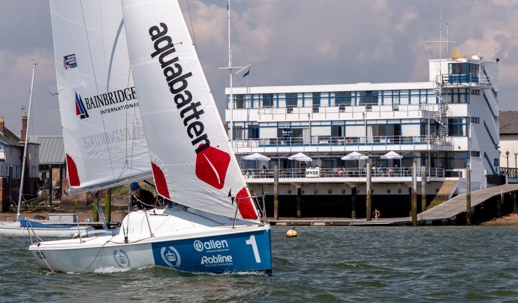 Shiny refurbished 707 keelboat sailing in front of Royal Corinthian Yacht Club's clubhouse on a sunny day, with umbrellas up on the balcony.