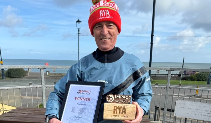 RYA Cymru Wales Impact Award winner Rob Davies with the wooden award and framed certificate, and wearing a blue drysuit and red woolly RYA CW hat on shore in the sunshine outside Llandudo SC with the sea in the background.