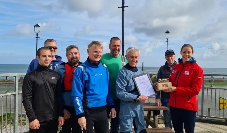 RYA Cymru Wales Performance Manager Sarah McGovern presents an Impact Award to Rob Davies surrounding by friends at Llandudno Sailing Club.