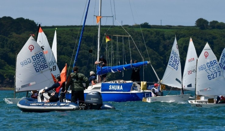 Handful of Optimists and an RS Tera gathered around blue yacht/committee boat in the sunshine at the Welsh Regional Junior Championships 2024.