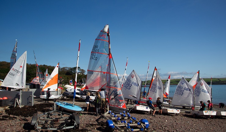 Variety of dinghies lined up on shore with colourful sails, waiting to launch.
