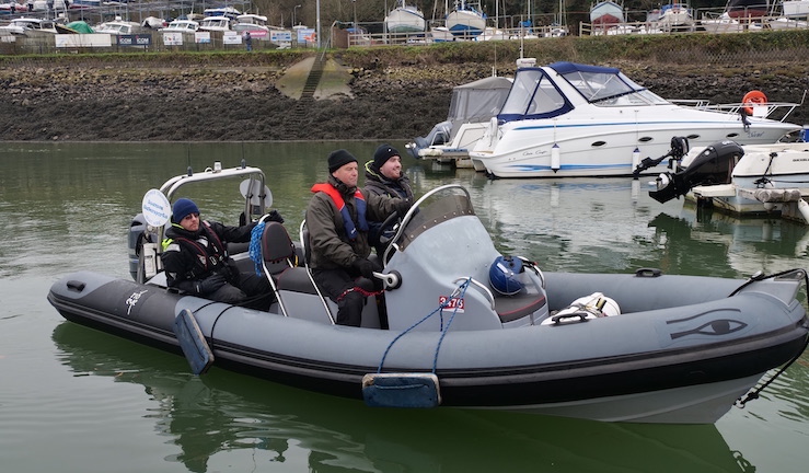 Grey powerboat with three people aboard in a harbour with a cruising powerboat also in the picture.
