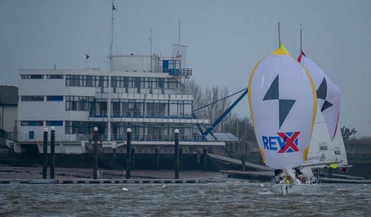 Two 707 keelboats match racing with spinnakers flying in front of the Royal Corinthian Yacht Club clubhouse on a grey and blustery day.