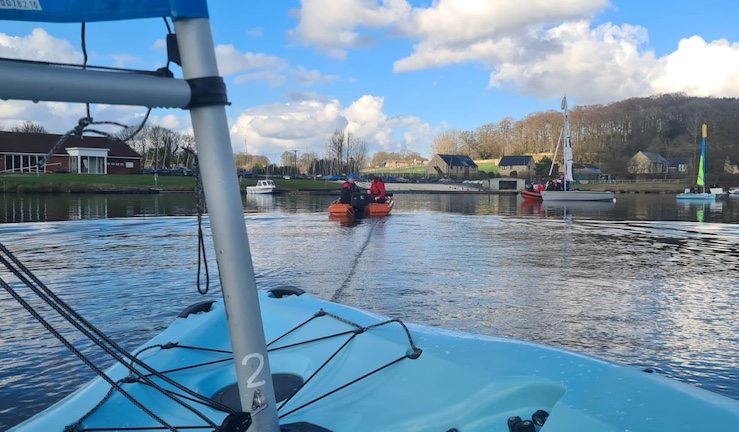 Pico dinghy's blue bow in the foreground with a tow rope to a powerboat in the distance with Ripon Sailing Club on shore.