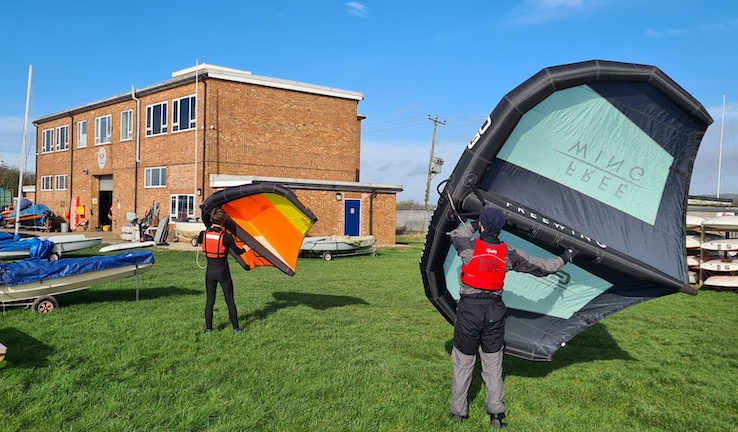Two people holding up wings on shore into the breeze with Welton Waters Adventure Centre redbrick building in the background.