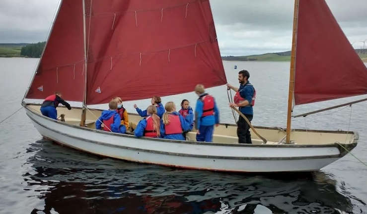 Aled Edwards standing up steering with the tiller at the back a boat with three red sails and half a dozen pupils on board, on a grey day with flat water. 