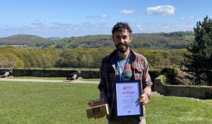 Outdoor education teacher Aled Edwards holding an RYA Impact Award certificate and wooden award with logo, against background landscape of lawn, rolling hills and blue sky.