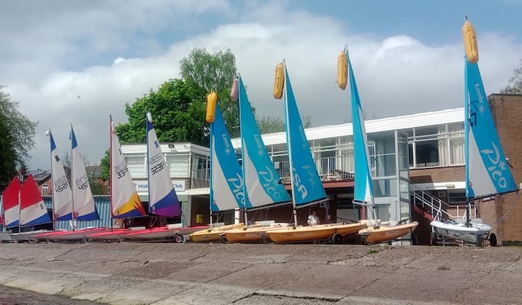 Five picos, four Toppers and two Optibat junior dinghies lined up with sails up on shore in front of the Midland SC / Sail Birmingham clubhouse.