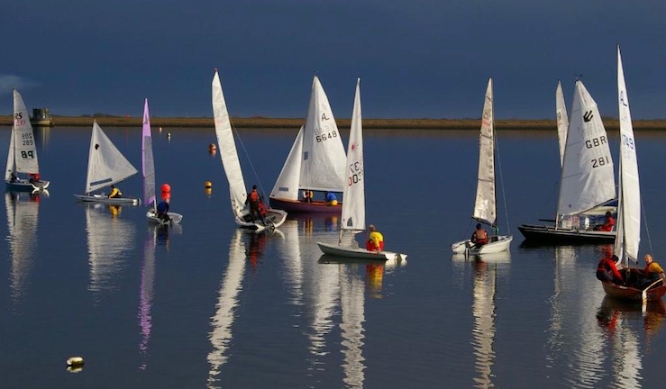 Fleet of variety of dinghies on flat water light wind day at Covenham SC with sunshine and reflections on the water.