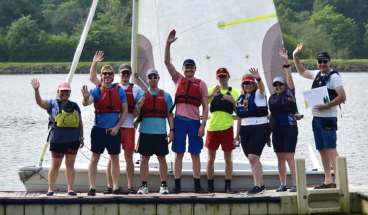 Nine people of all ages waving on the pontoon at Trimpley SC with a boat behind them with its white sail as a backdrop.