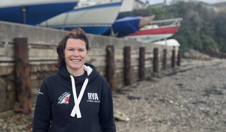 RYA Cymru Wales Club Development Officer Hester Walker in blue Welsh Sailing branded hoody on a beach pictured in front of keelboats in a boat park.