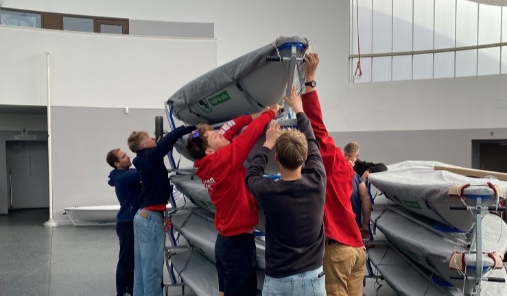 Group of Welsh youth squad sailors helping to unload a new ILCA from a multi-stacker in the Plas Heli hangar.