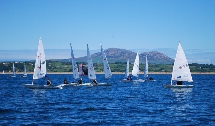 ILCA dinghies sailing with blue seas and skies and mountains on the horizon.