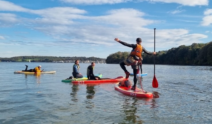 People taking part in a SUP session with an instructor demonstrating standing on one leg and balancing holding out arm and paddle.