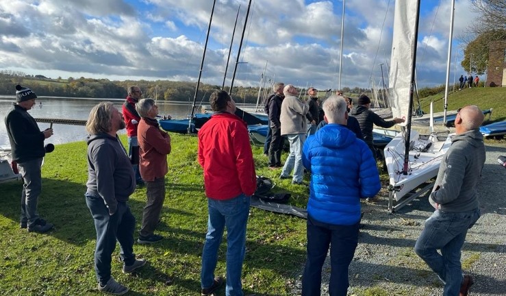 Group of instructors stood around looking at a rigged boat with sail up on shore during a 'Ben in a Boat' upskilling visit to Chelmarsh Sailing Club.