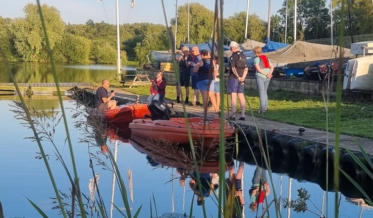 Regional manager Ben Hodgson in an orange powerboat alongside and chatting to a shoreside group at Syston Sailing Club during a 'Ben in a Boat' upskilling visit.