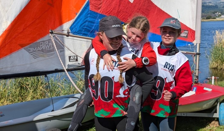 Three smiley young Welsh sailors in Dragon bibs and stood next to a Topper with red white and blue sail at the RYA CW OnBoard Festival at Bala, 2024  