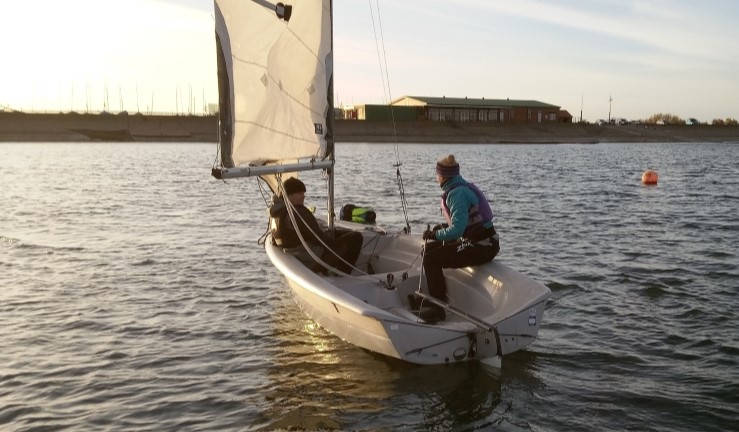 A dinghy being sailed by two people at sunset towards the clubhouse at Covenham Sailing Club.