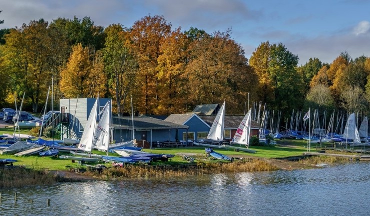 View of Hollowell SC clubhouse from the water with boats rigged on the shore and a backdrop of trees with leaves turning from green to autumn colours.