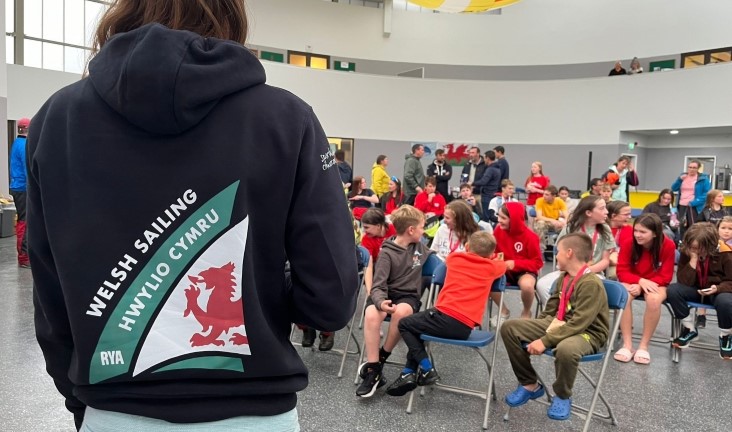 Back view of RYA Cymru Wales staff member sporting new branded hoodie with Welsh Sailing logo, talking to crowd of junior sailors inside at Plas Heli.  