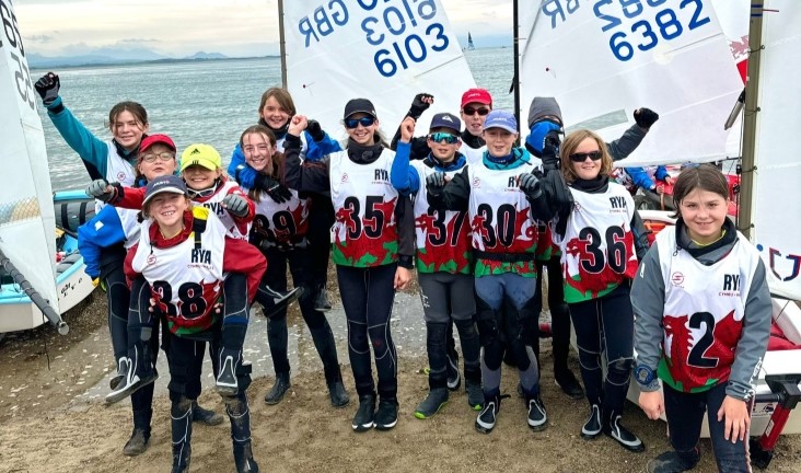 A dozen smiley Oppie sailors sporting Welsh dragon bibs on the beach next to their boats at Welsh Youth & Junior Championships 2024.
