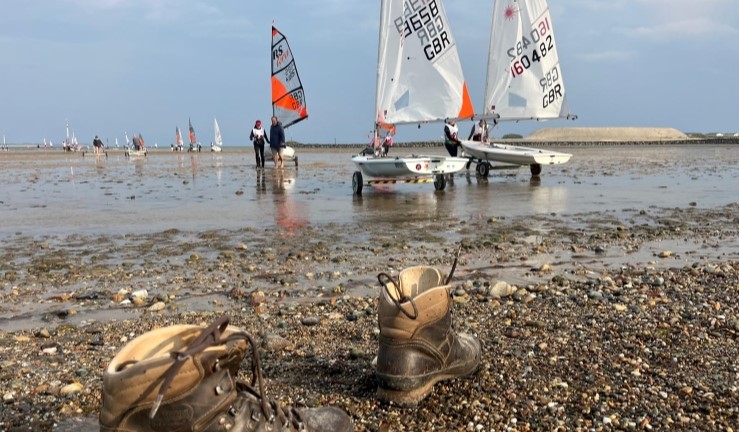 Walking boots left on shore with view over the beach, tide well out, with boats being walked over the wet sand. 