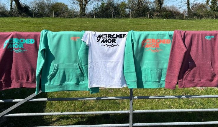 Five T-shirt/hoody tops in white/green/red with Merched Y Mor branding hanging over a farm gate in the sunshine.