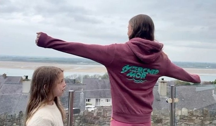 Woman looking out to sea with arms outstretched and hoody branded Merched Y Mor with young girl alongside.