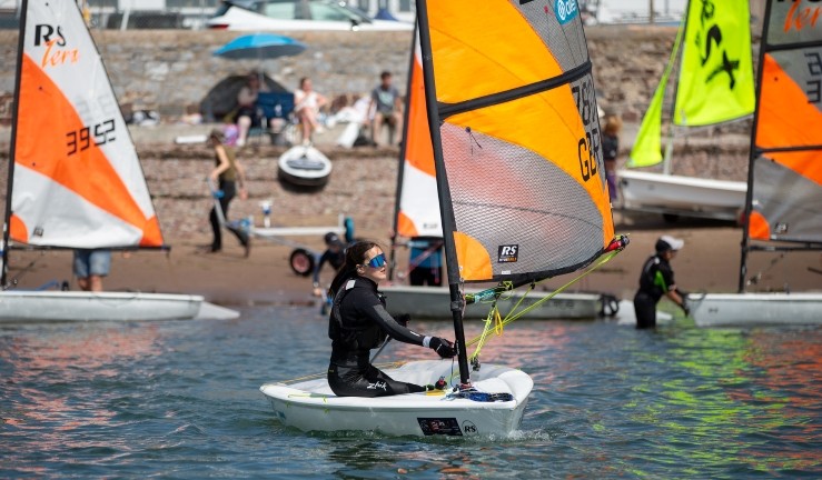 A view from the water of a girl launching from the beach at Dale for a Dragon Series event wearing sunglasses and sailing an RYA Tera with an orange sail, with other children also getting their boats ready to launch.