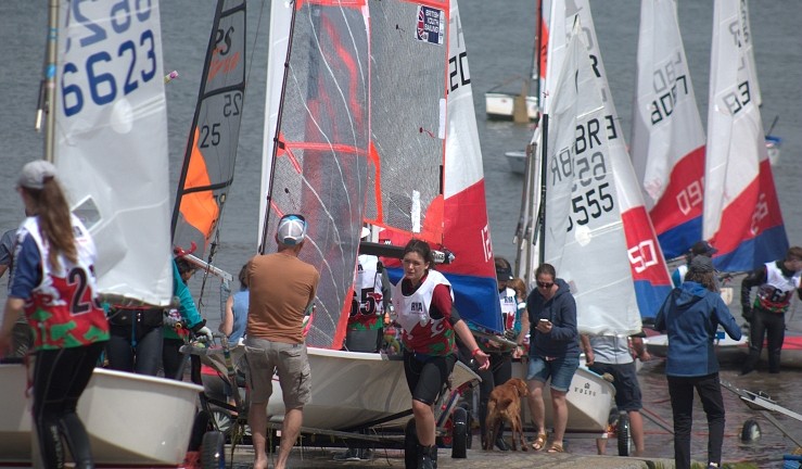Variety of colourful youth and junior dinghies being pulled up a beach by sailors and parents, with classes including Optimist, Topper, RS Tera and 29ers. 