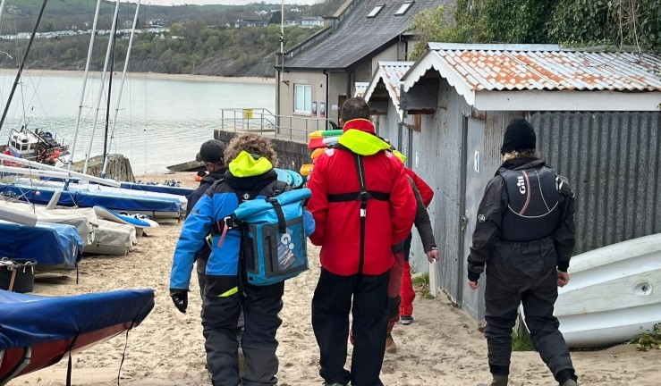 Three people wearing sailing kit/coats/life jackets and carrying rucksacks walking through a boat park towards the shore at an RYA CW Regional Traning Day 2023, Cardigan Bay Watersports.
