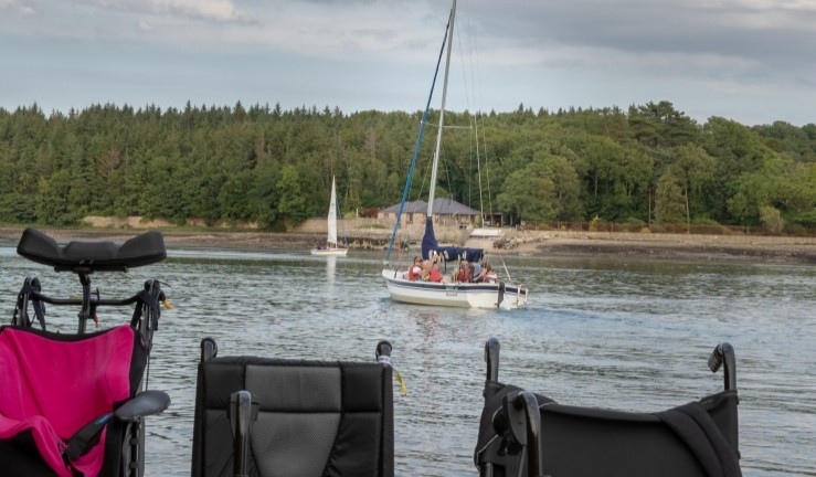 Wheelchairs left on shore with a view of a SEAS Sailability group aboard a yacht enjoying the freedom of being on the water.