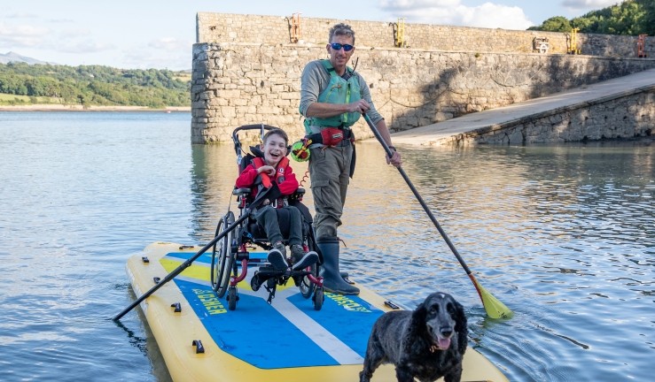 A child in a wheelchair all smiles aboard a paddleboard with a dog and SEAS Sailability volunteer.