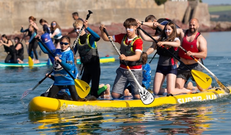 Half a dozen people of all ages having fun in the sun paddling a giant yellow paddleboard.