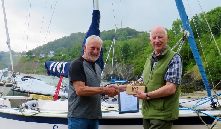 Cardigan Bay Watersports principal Crawford McLeary presents an RYA Cymru Wales Impact Award to Stephen Wood, onshore with keelboat in background.