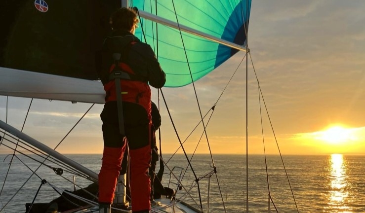Bow of BSKA yacht on offshore weekend with sailor and green spinnaker sailing towards a sunset.