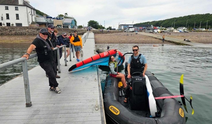 Pontoon at Dale with shoreline in the background and group of people smiling for the camera and RIB alongside with two people aboard and winging kit.