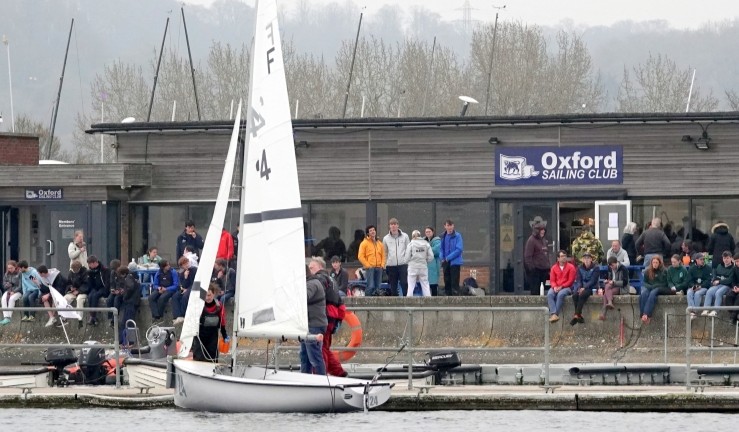 View from the water of spectators in front of Oxford Sailing Club at the RYA/UKTRA National Team Racing Championship 2025 with a Firefly dinghy team changeover on the pontoon.