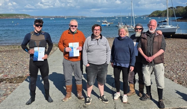 Half a dozen newly qualified RYA Safety Boat holders on a slipway with the sea behind them after completing their certificates as part of the RYA CW Big Day Out 2025 in Pembrokeshire. 