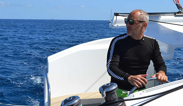 Man at helm of catamaran in the Mediterranean. Winches in foreground, boom in background. Blue skies, sunny day.  