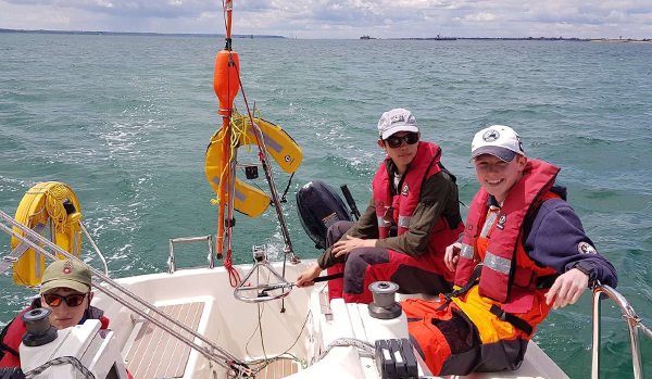 three young men in yacht cockpit sailing. All wearing caps and red lifejackets, looking at the camera and smiling.
