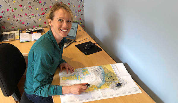 Woman doing RYA navigation course is sat at a desk with a laptop, charts and a portland plotter. She is looking at the camera and smiling. 