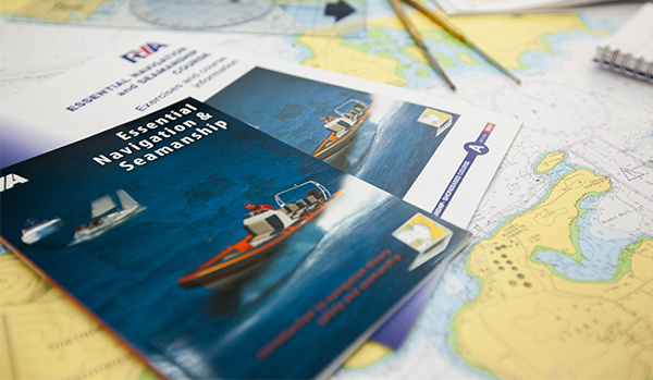 RYA Essential Navigation and Seamanship course booklets on desk with charts, notebook, plotter and dividers.